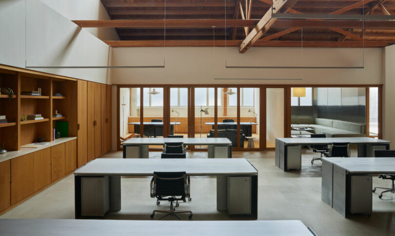 An open office space with wooden walls and beams, multiple workstations with chairs, and a glass partition separating another room. Desks have no personal items. The area is well-lit with natural light.