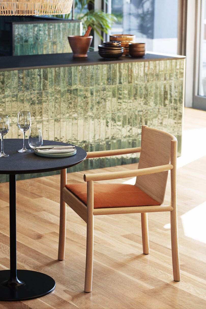 A Kata Chair with an orange cushion is placed next to a small black table set with three wine glasses, a white plate, and cutlery. A green tile backsplash and wooden floor are visible in the background.