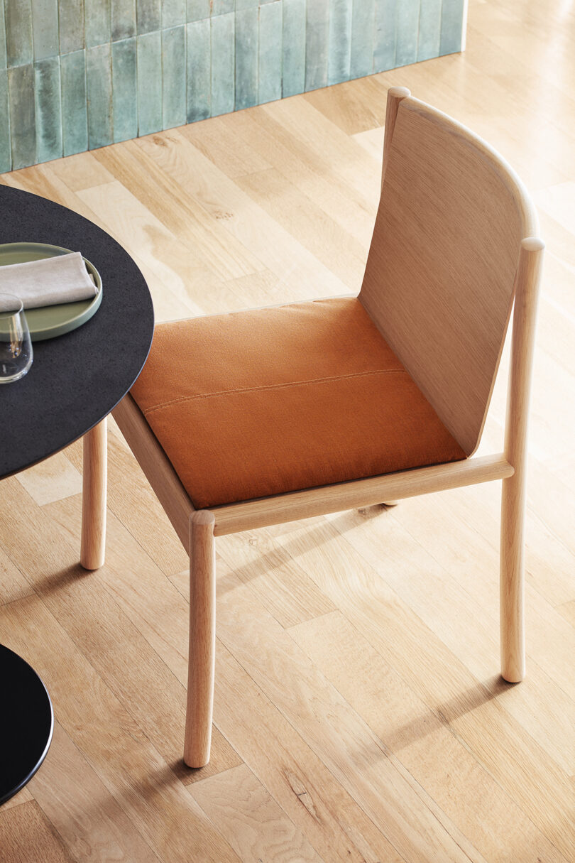 A Kata Chair with an orange seat cushion next to a round black table set with plates and a napkin. The floor is made of light wood, and a textured green wall is in the background.