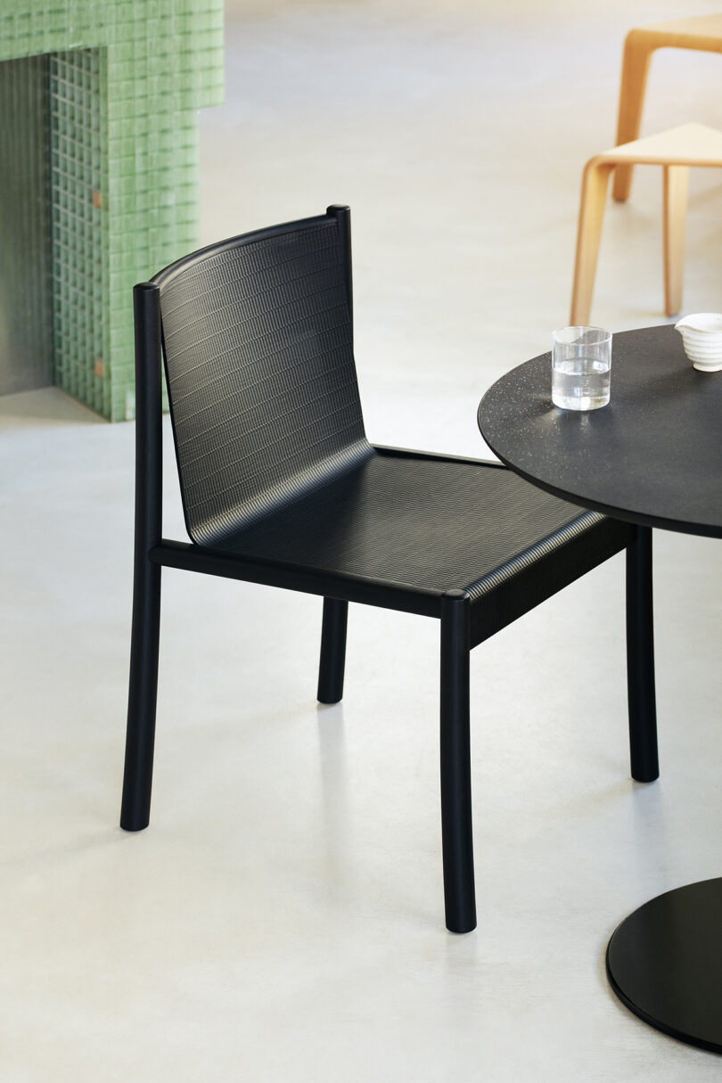 A black Kata Chair next to a round black table with a glass of water and two white cups on it, all placed in a modern, minimalist interior space.