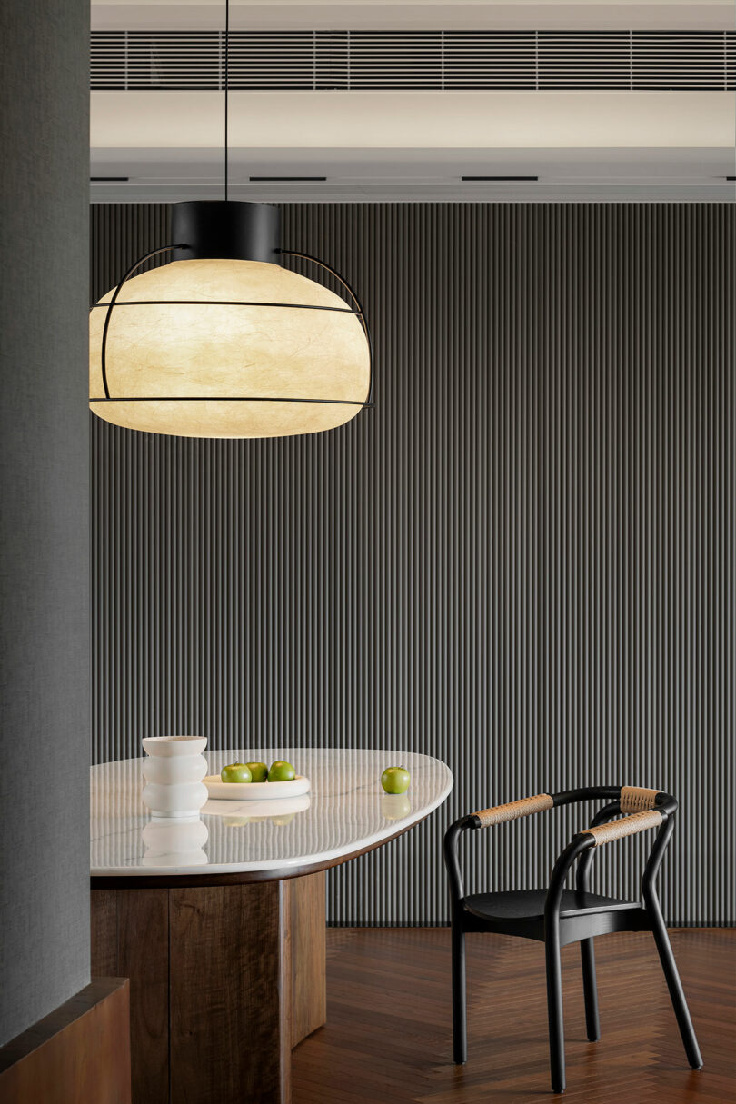 Modern dining area with a dark vertical striped wall, a wooden table set with white bowls and green apples, a black chair with a wicker seat, and a large round pendant light above.