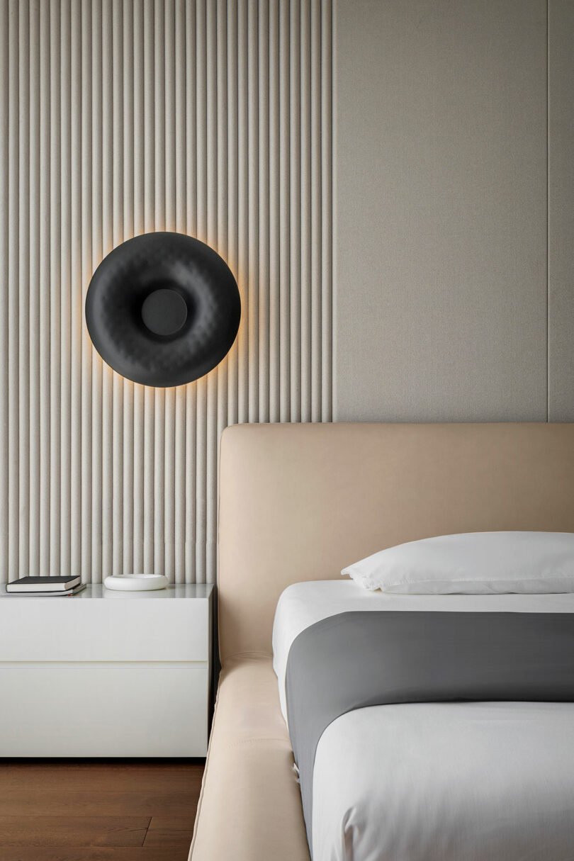 Minimalist bedroom interior featuring a beige upholstered bed, white bedside table with books, and a round black wall light against a textured panel wall.