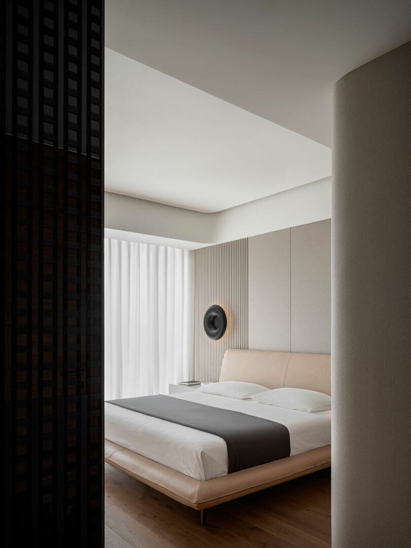 Modern bedroom with a low-profile bed, beige headboard, and grey bedding. The room features minimalist decor, light wood flooring, a circular wall art piece, and floor-to-ceiling white curtains.