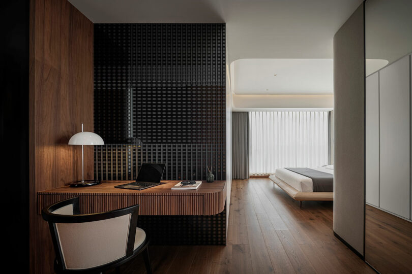 Modern hotel room with a sleek desk setup featuring a laptop, lamp, and chair. The room includes a bed, large window with curtains, and wooden flooring.