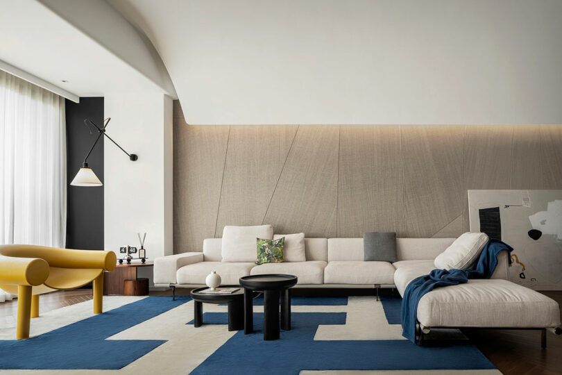 Modern living room with a white sectional sofa, yellow armchair, blue and white geometric rug, and black coffee tables. There is a minimalist wall-mounted light fixture and abstract wall decor.