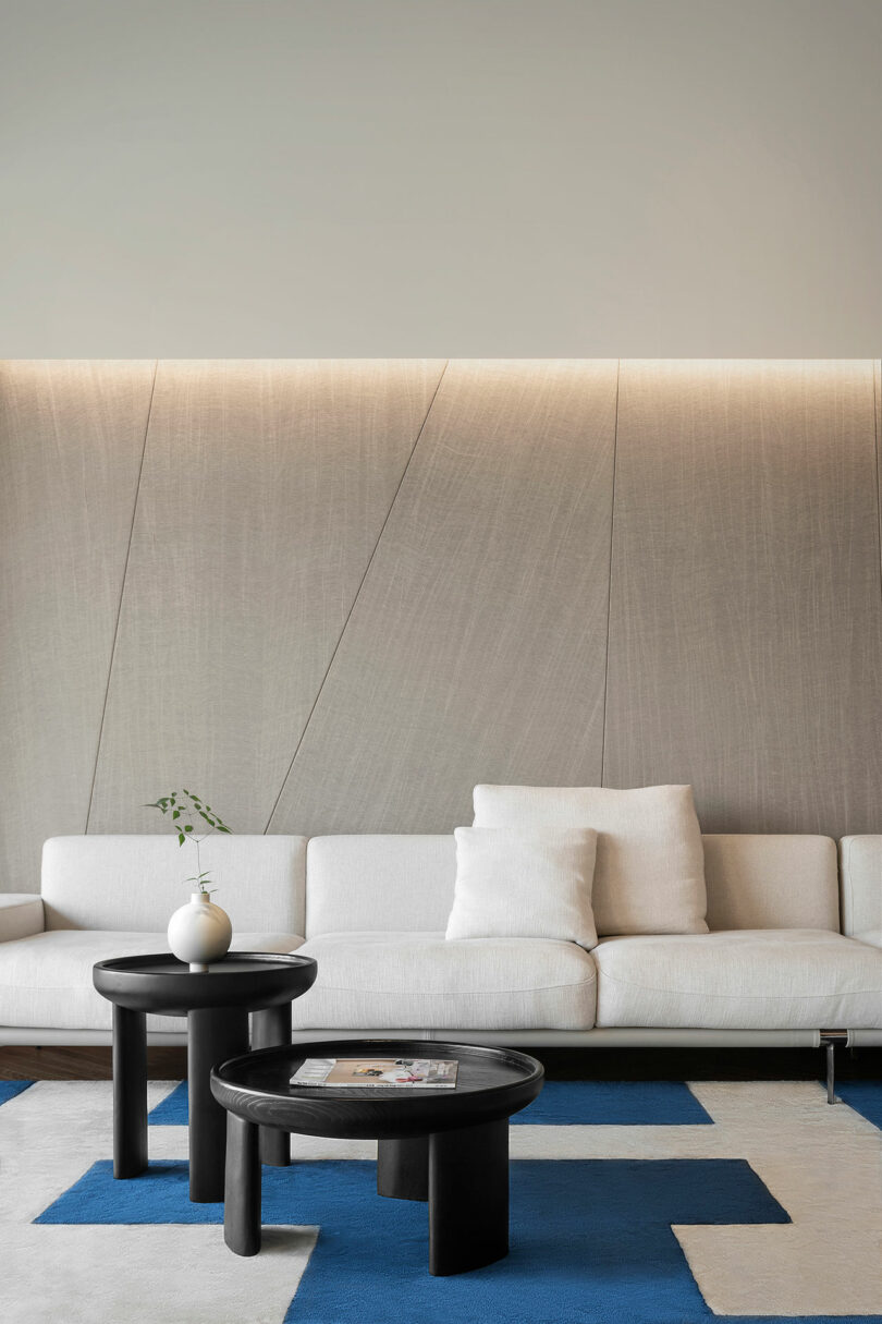 A modern living room features a white sofa with two black coffee tables, a vase with a sprig of greenery, and a minimalist wall with soft lighting. Blue and white geometric rug on the floor.