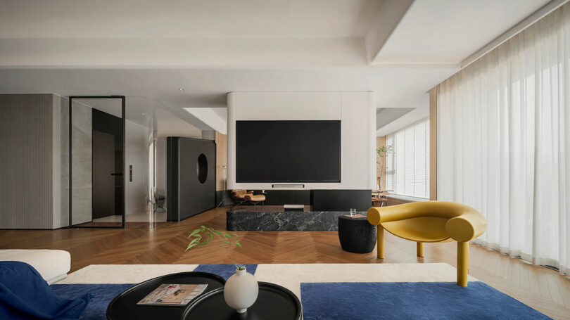 A modern living room with a large TV mounted on a wall, a yellow chair, two round tables, and a potted plant. The room features wooden flooring and large windows with sheer curtains.