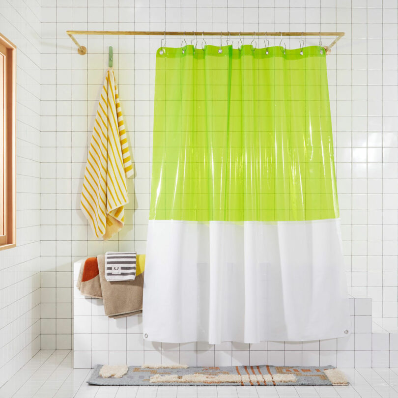 A bathroom featuring a white-tiled wall, a transparent green and white shower curtain, striped yellow towel, and multicolored towels hanging on hooks like a colorful lineup of well-behaved brats, with a bath mat on the floor.