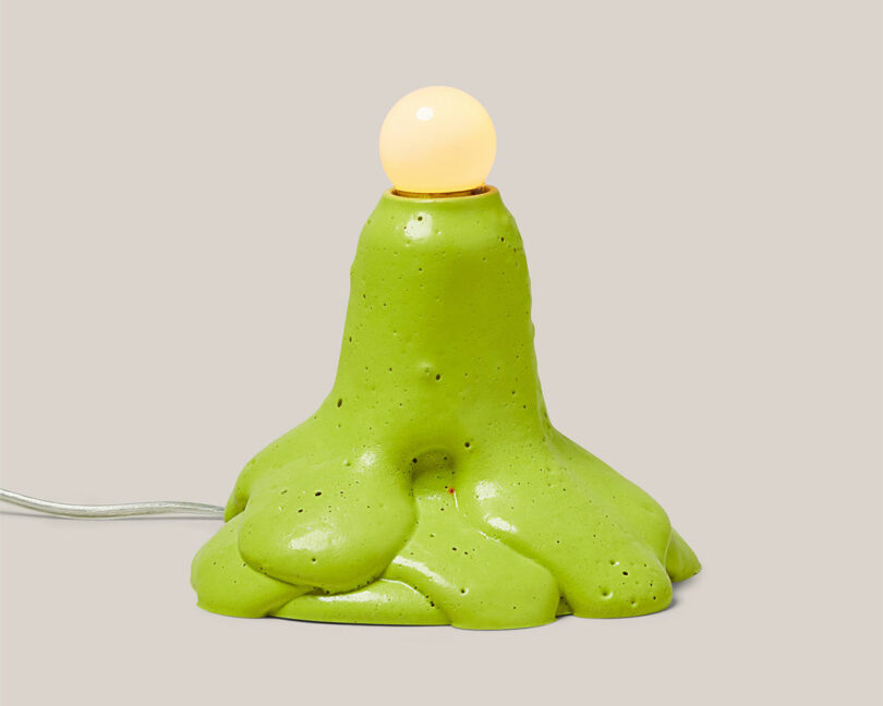 A green, organic-shaped lamp base resembling melting clay and embodying the whimsical spirit of a brat, topped with a lit round bulb.