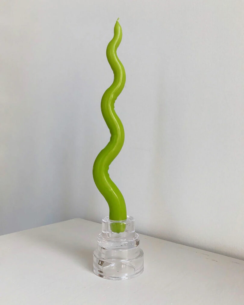 A brat-green twisted candle in a clear holder stands on a white surface against a plain white background.