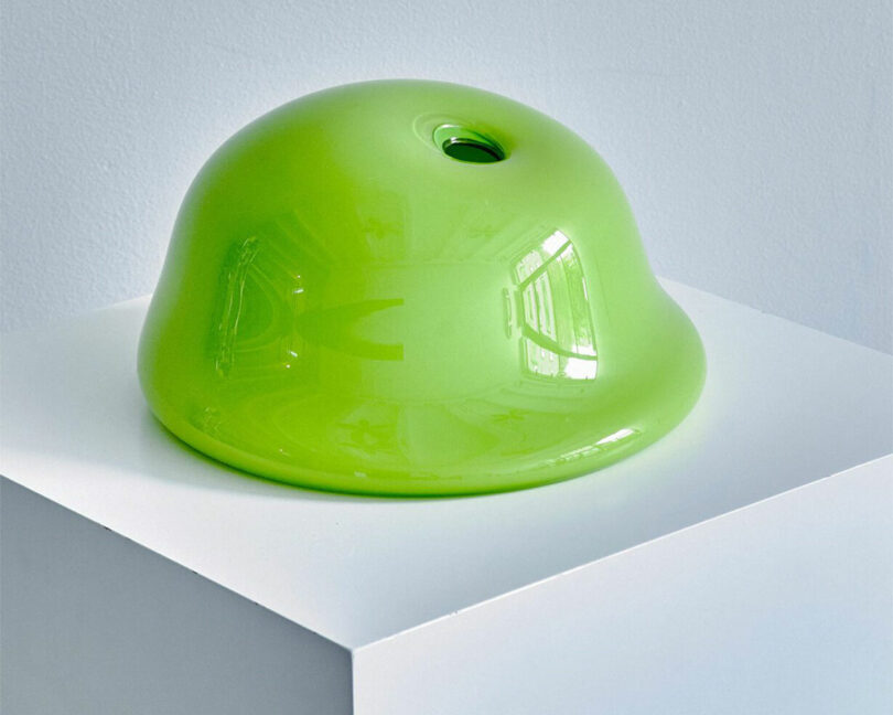 A shiny, lime green, dome-shaped object with a small hole at the top is displayed on a white pedestal, much like a brat showing off against a light grey background.