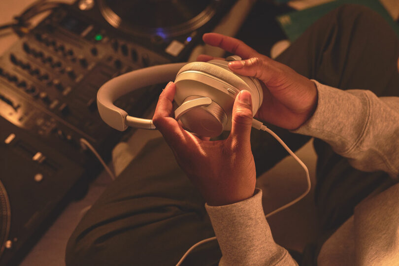 Person adjusting Melomania P100 white over-ear headphones with audio equipment in the background.