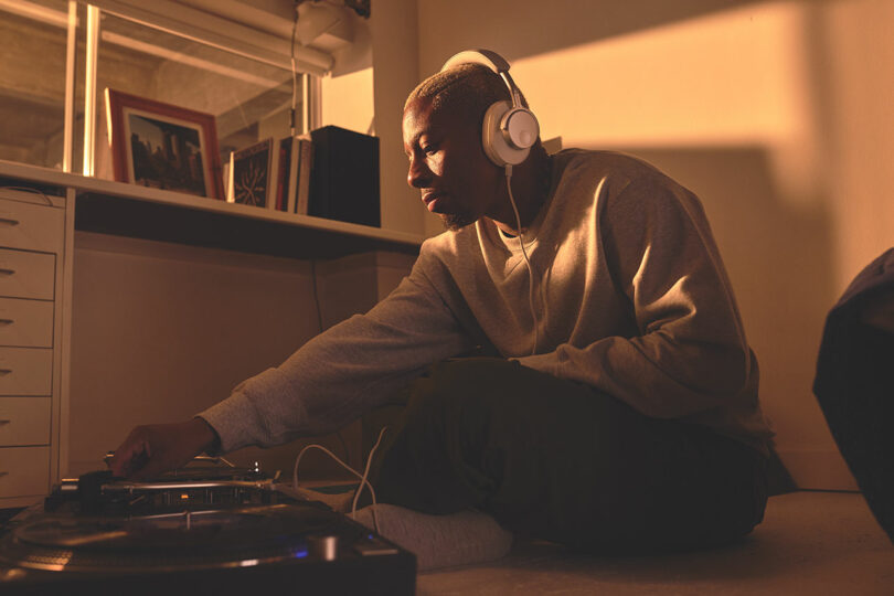 A person sitting on the floor wearing Melomania P100 headphones, adjusting controls on a DJ turntable in a room with a window and various framed photos on a shelf.