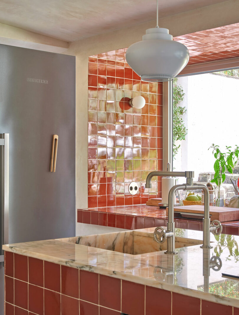 Modern kitchen with red tiled counters and backsplash, marble island with a sink and a hanging light fixture. Stainless steel refrigerator is visible on the left.