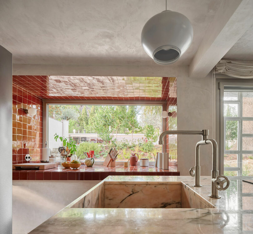 Modern kitchen interior with a large marble sink, red tiled backsplash, and a view of a green garden through the window. A pendant light hangs above the sink area.