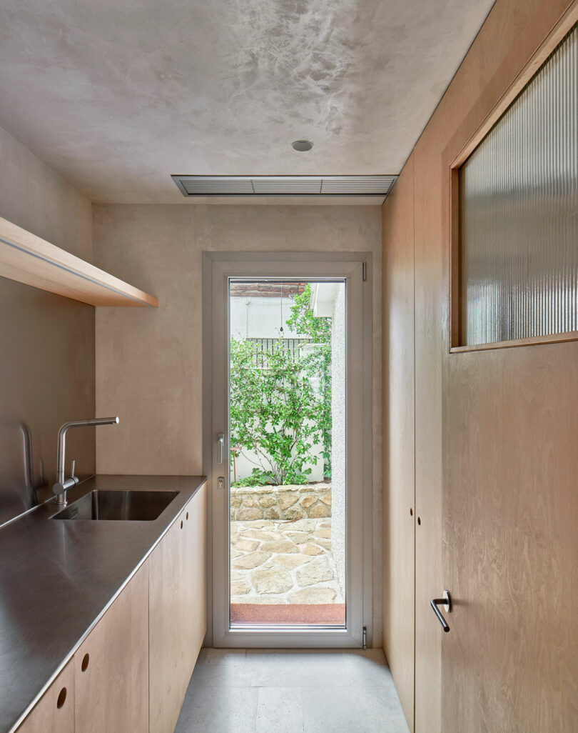 A small, minimalist kitchen with a stainless steel countertop, dual sinks, wooden cabinets, and a glass door leading to a garden outside.