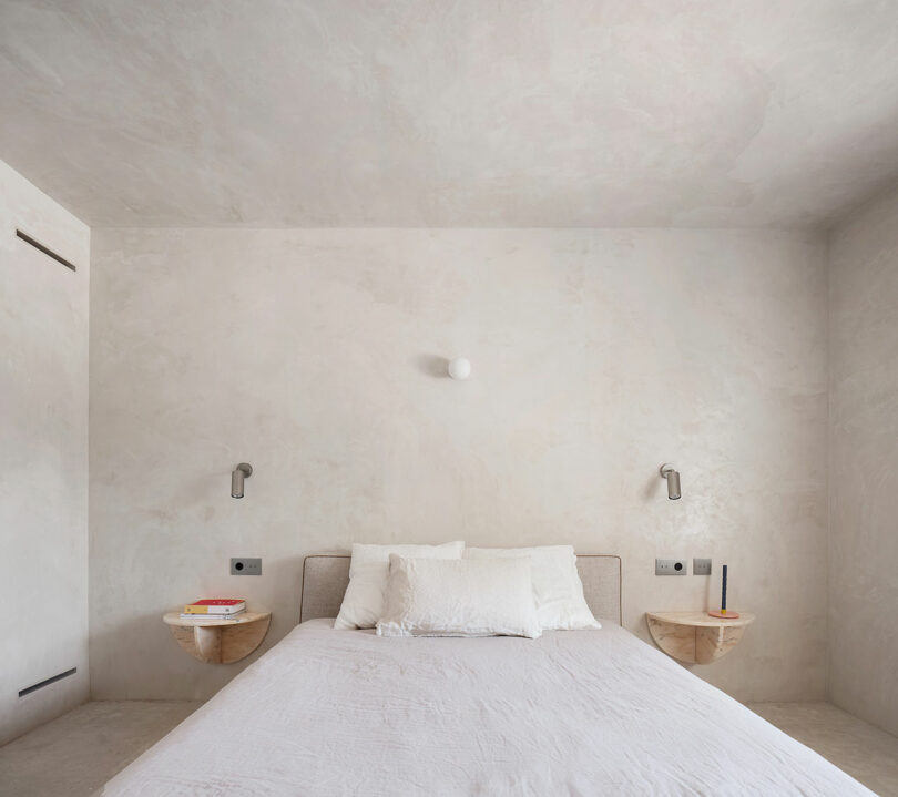 A minimalist bedroom with light-colored walls features a neatly made double bed, two wall-mounted nightstands with books and a candle, and two modern wall lamps on either side.