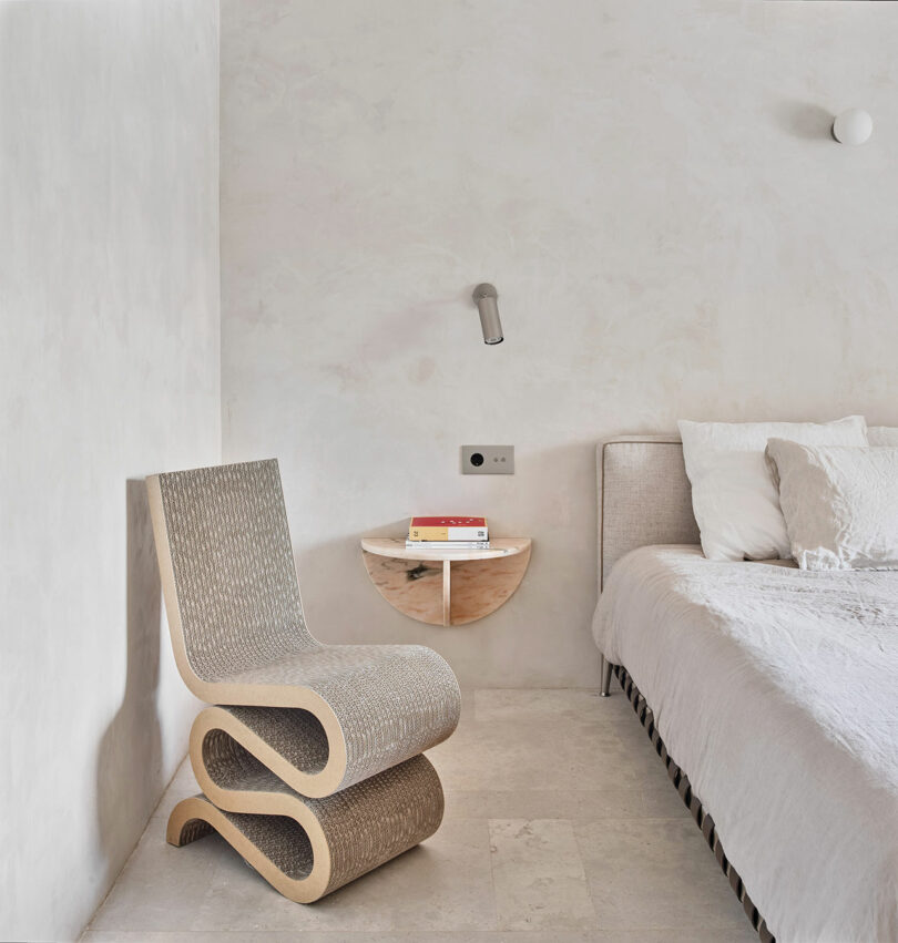 A minimalist bedroom with a modern S-shaped chair, textured walls, a circular wooden bedside table with books, and a bed with white linens.