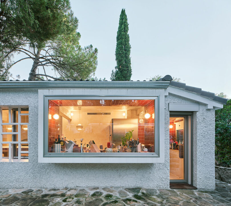 A view of a modern kitchen through a large window in a stucco house with a pathway outside. The kitchen is brightly lit with hanging lights and houses various appliances and kitchenware.