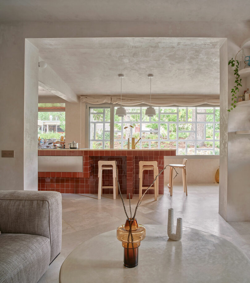 A modern living space with an open plan kitchen featuring a red-tiled island, white bar stools, a beige sofa, a sleek coffee table, and a large window providing natural light.