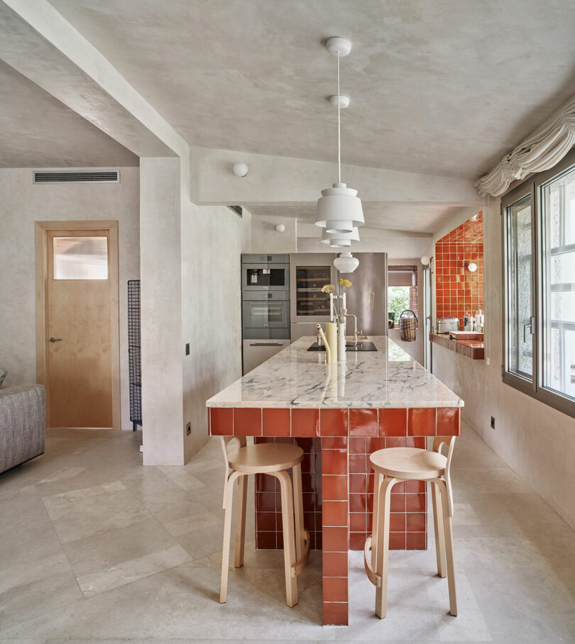 Modern kitchen with a long marble-topped island, red tiled base, two wooden stools, pendant lights, and stainless steel appliances. Large windows provide ample natural light.