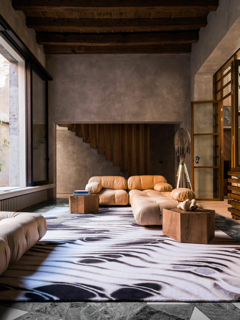 A modern living room with a wooden ceiling and staircase, cream-colored plush sofa set, wooden tables, and an abstract black-and-white patterned rug. Natural light enters through a large window, highlighting a stunning Dale Chihuly glass sculpture as the room's centerpiece.