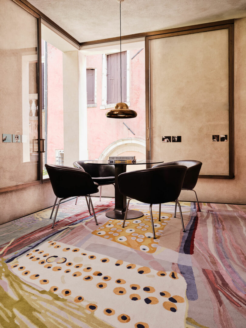 A small dining area with a round table, four black chairs, and a hanging pendant light. The floor features a colorful patterned rug, reminiscent of Dale Chihuly's vibrant designs. Large windows offer a view of an exterior pinkish wall and arched window.