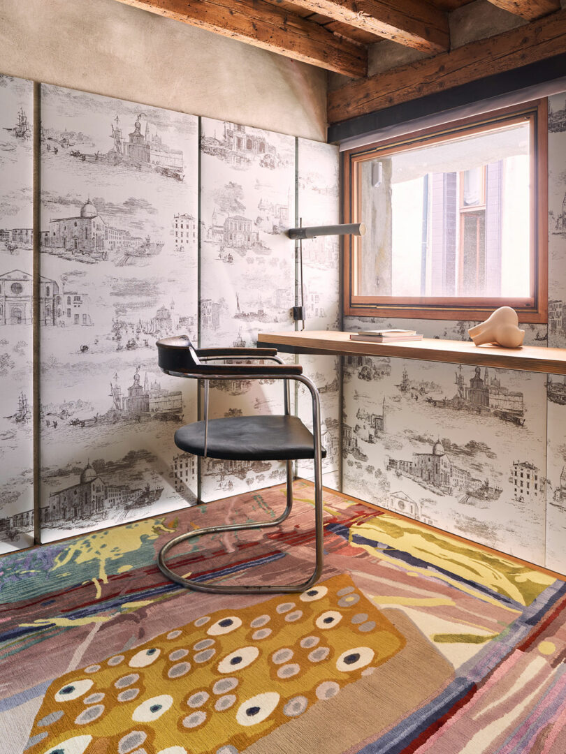 A modern study with a patterned wall, a wooden desk beneath a window, a black chair, and colorful rugs on the floor features vibrant Dale Chihuly glass art pieces.