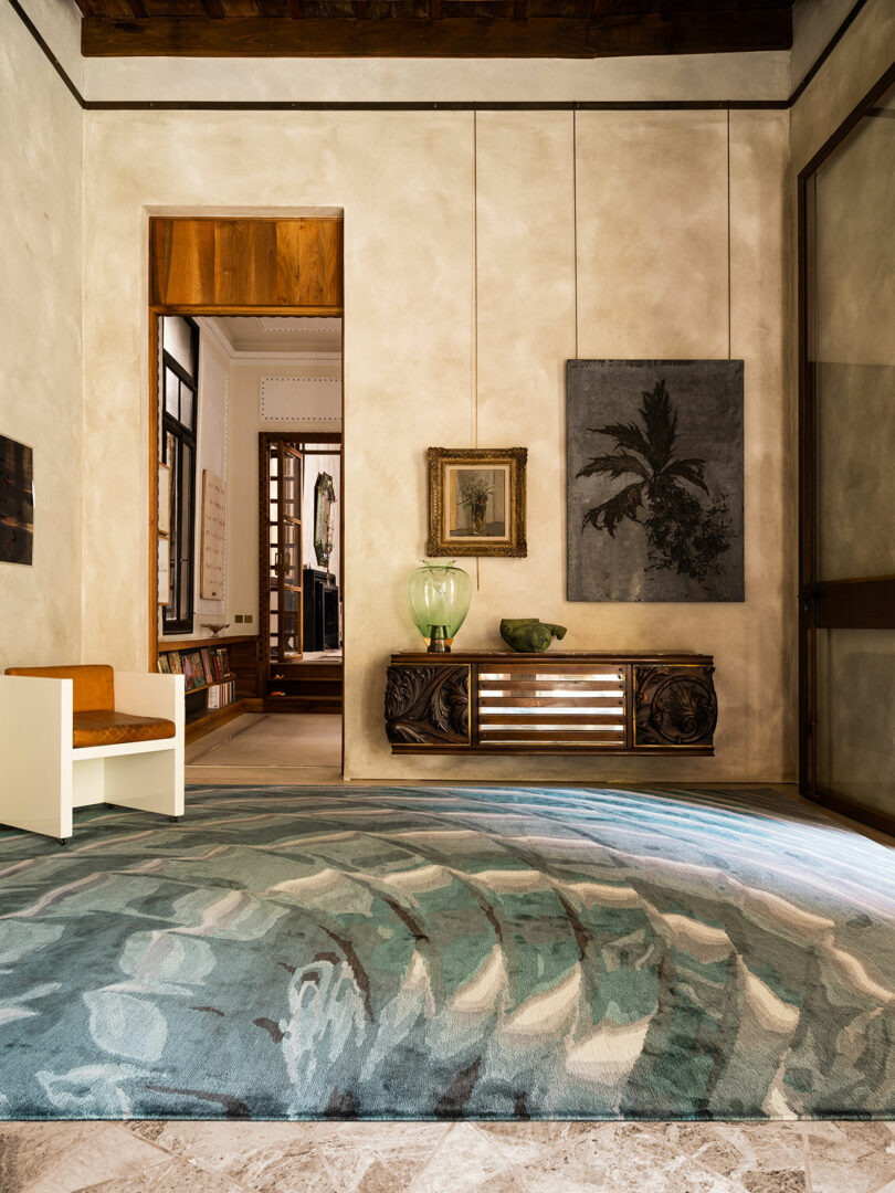 A well-lit room features a patterned rug, a white and brown chair, wall art reminiscent of Dale Chihuly's vibrant glasswork, and a wooden console table holding decor items like a lamp and a bowl. An open doorway leads to another room with bookshelves.