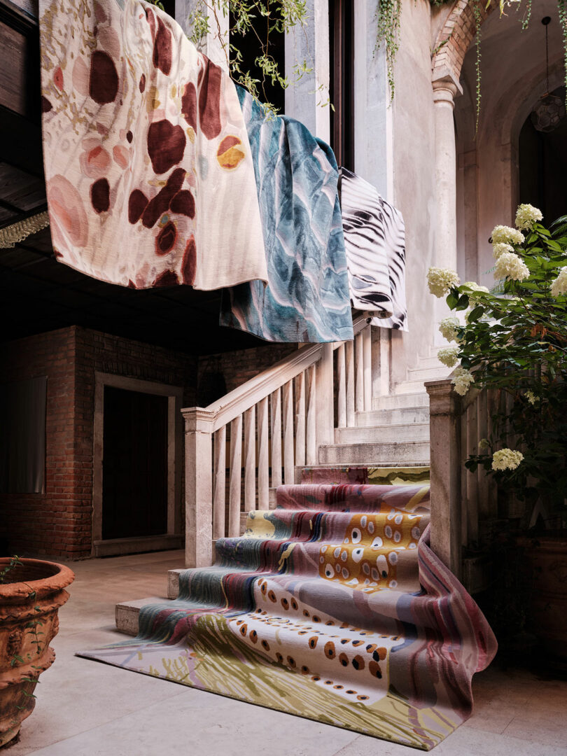 Colorful textiles are draped over a railing above a stone staircase adorned with a patterned rug; plants are visible on both sides, evoking the vibrant, artistic touch reminiscent of Dale Chihuly.