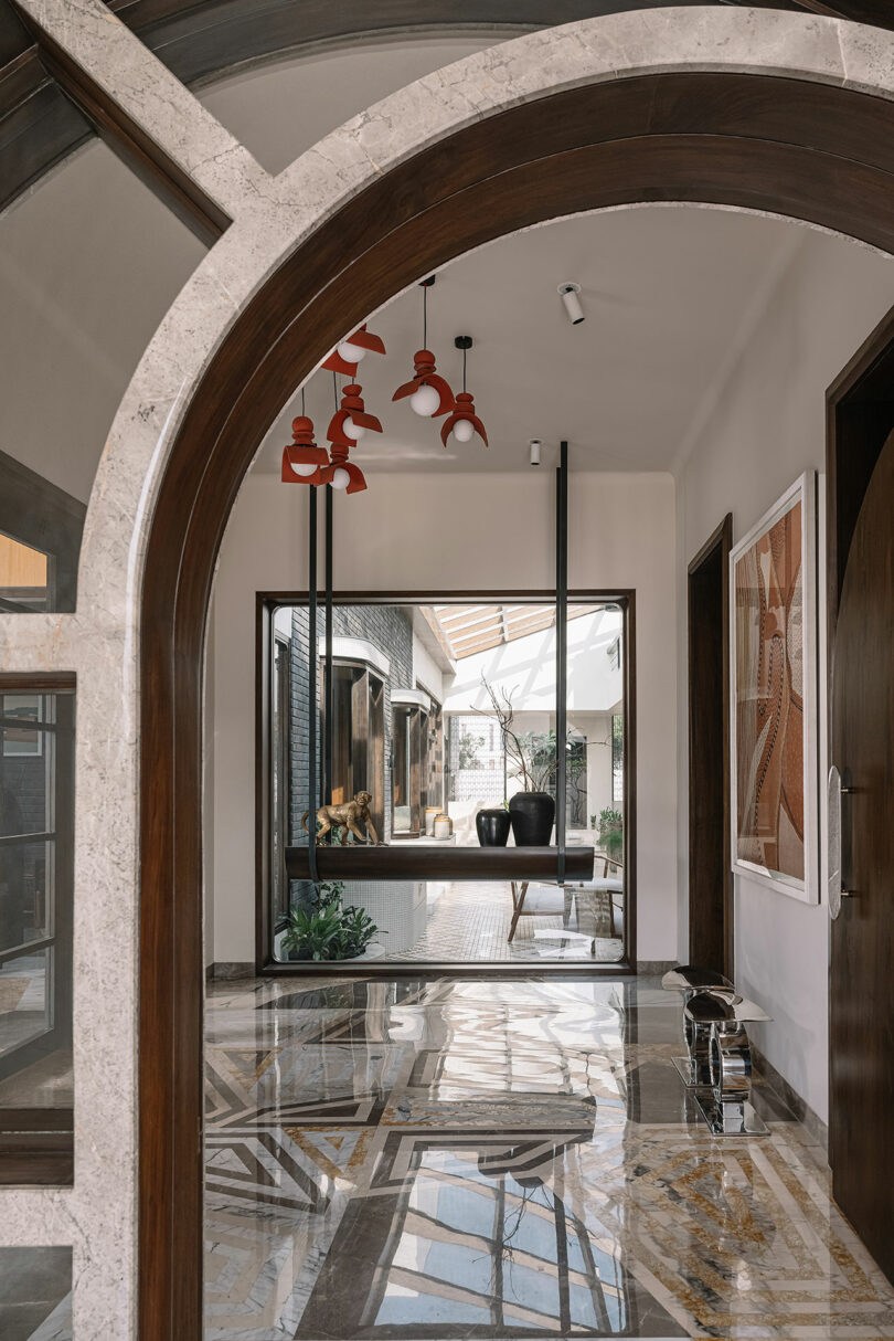 Arched hallway with marble flooring, modern red light fixtures, and large window overlooking an outdoor area with seating. Artwork hangs on the walls and light reflects off the shiny floor.