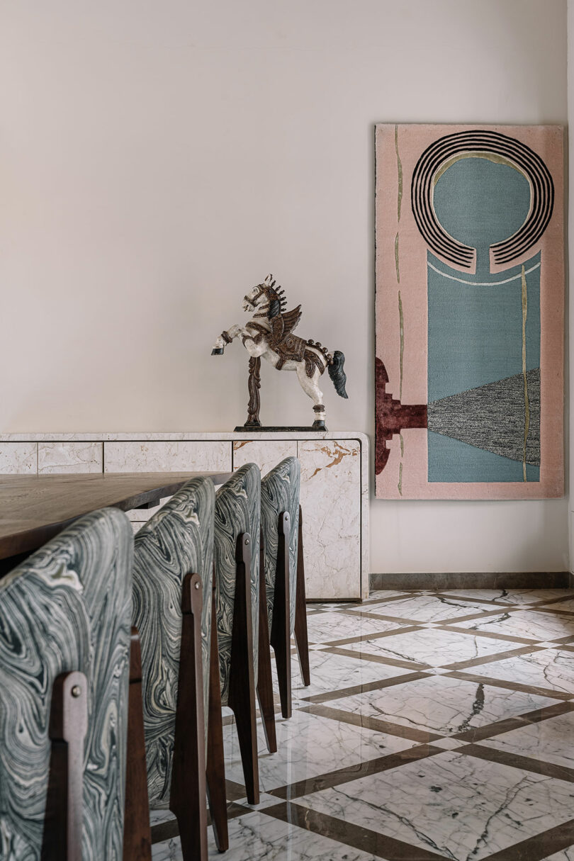 A dining area with a marble table and green upholstered chairs. A wooden horse statue on a marble sideboard and a modern abstract art piece decorate the wall above it.