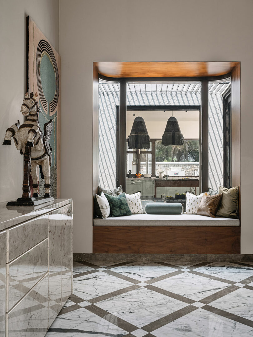 A cozy nook with cushioned seating and throw pillows, adjacent to a well-lit space with large windows and hanging lamps. A marble-tiled floor and a sideboard with decorative items are also visible.