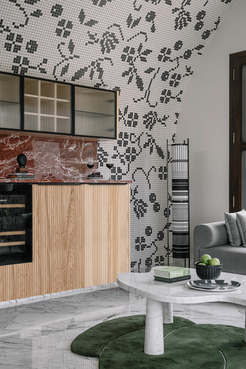 A modern kitchen with floral mosaic tiles on the wall, wooden cabinets, a striped towel rack, a marbled countertop, a gray sofa, and a round white coffee table on a green rug with books and fruits.