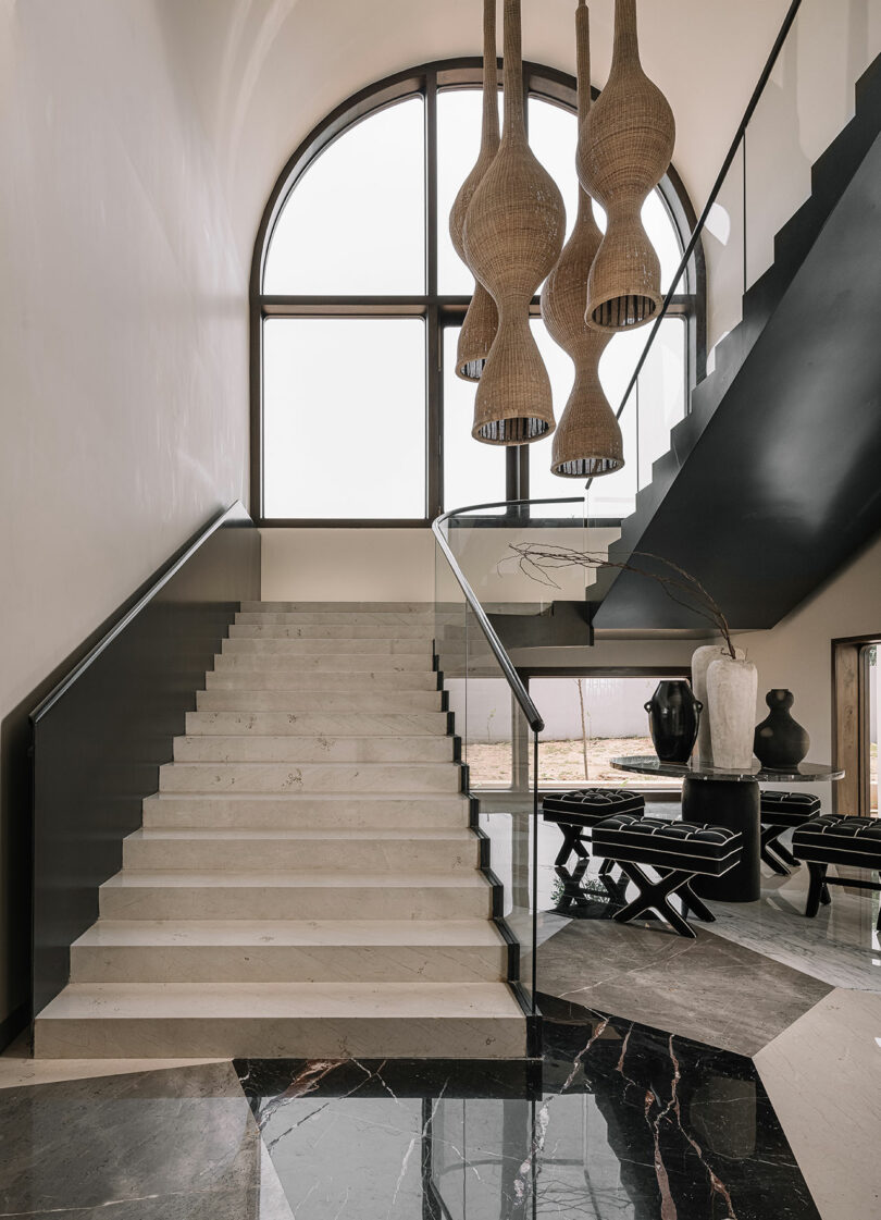 A modern interior with a staircase featuring stone steps, black railings, large arched window, woven pendant lights, and black and white vases on a table in the background.