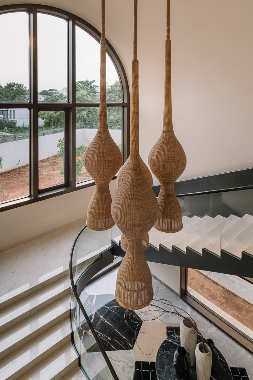 A modern interior features large wicker pendant lights hanging above a curved staircase with black and white marble steps, next to a large arched window overlooking a garden.