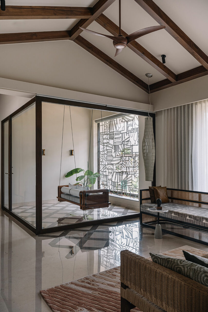 Modern living room with a hanging chair, glass walls, wooden beams, a ceiling fan, and a potted plant. Neutral tones and minimalistic decor create a spacious and calm atmosphere.