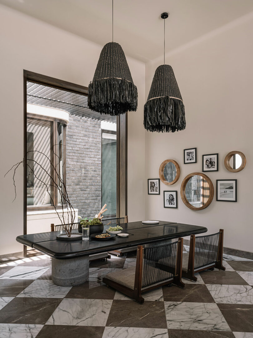 Modern dining room with a dark wooden table, low wooden chairs, and two large black pendant lights. A wall features round mirrors and framed photos. Large window opens to brick exterior.