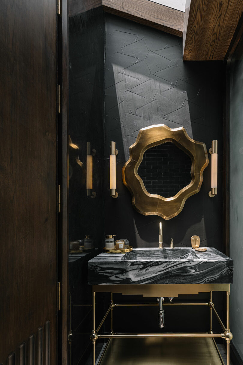 Modern bathroom with a black marble sink, brass fixtures, a wavy-edged gold-framed mirror, dark textured walls, and two vertical light sconces.