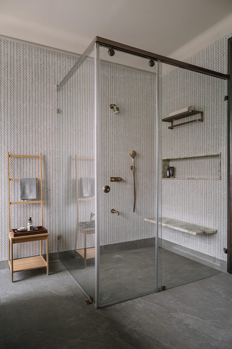 A modern bathroom features a glass shower enclosure, patterned wallpaper, and a wooden towel rack with shelves.