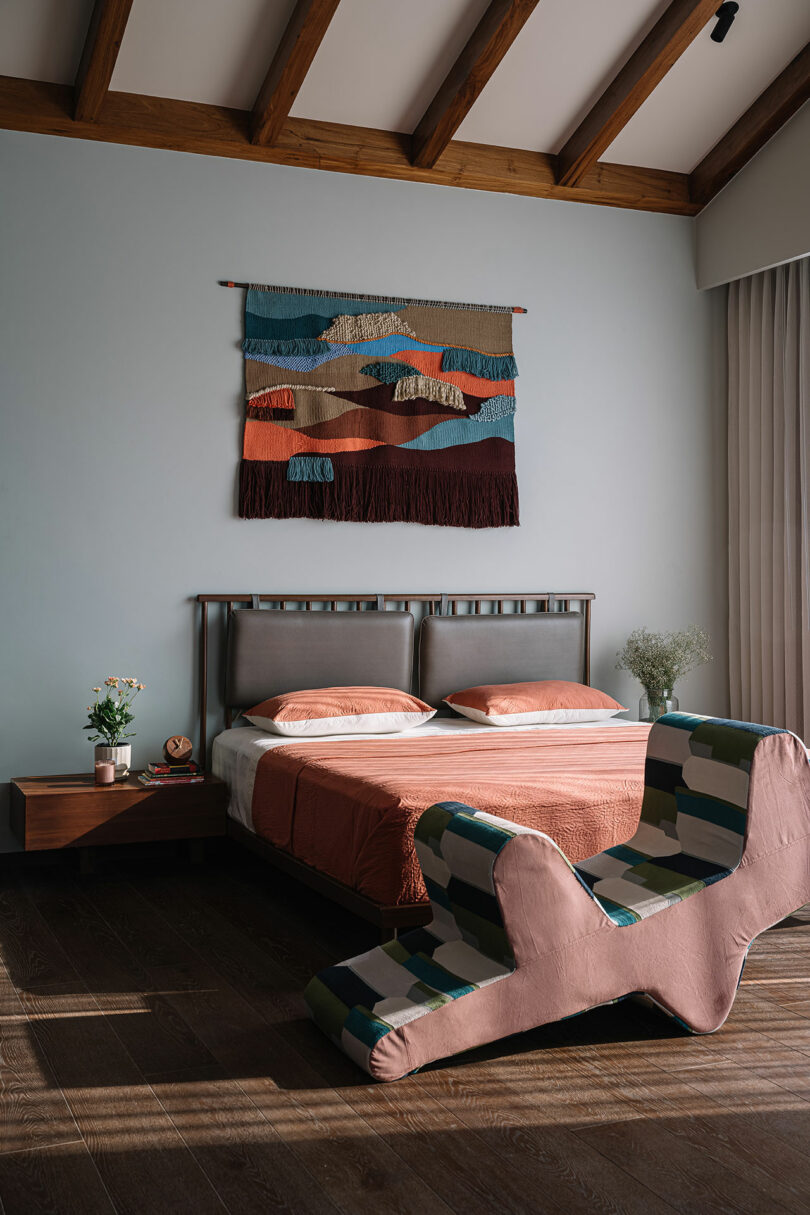 A bedroom with a double bed, rust-colored bedding, a wooden bedside table with flowers, a multicolored wall hanging, and a modern chair with a geometric design.