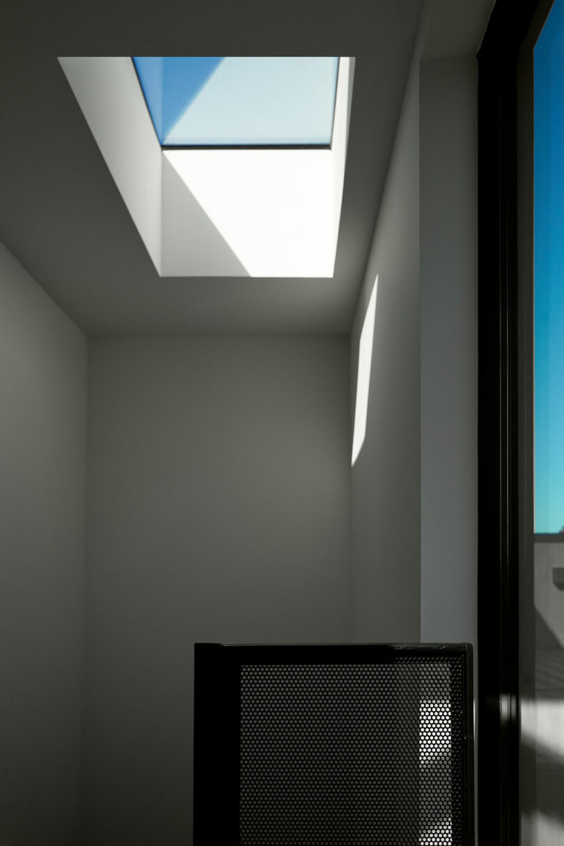 A minimalist room with a square skylight casting shadows on white walls and part of a mesh chair in the foreground.