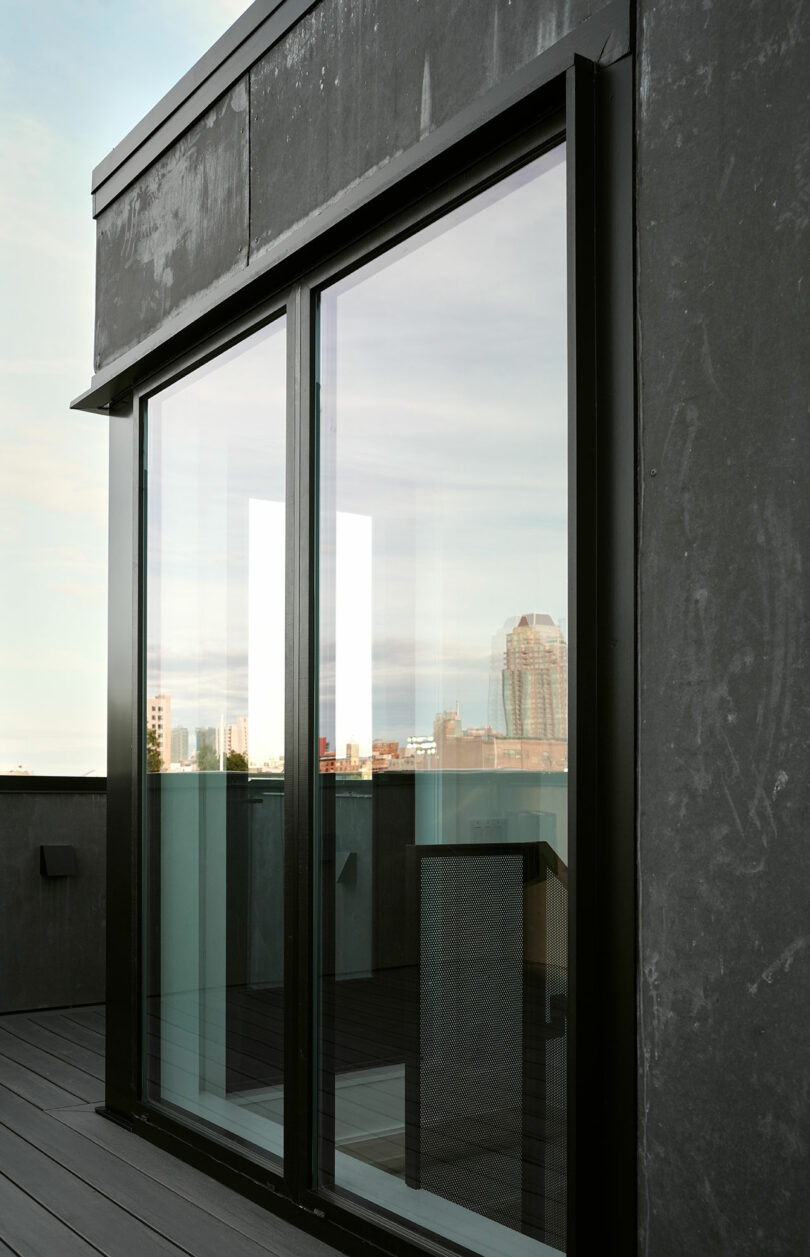 A modern sliding glass door on a building's rooftop, reflecting the city skyline in the background. The exterior is composed of sleek, dark panels.