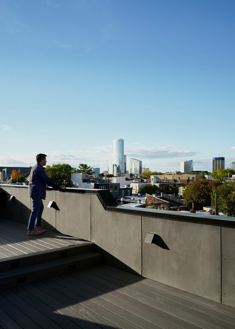 A person stands on an elevated outdoor area with views of a city skyline under a clear blue sky.