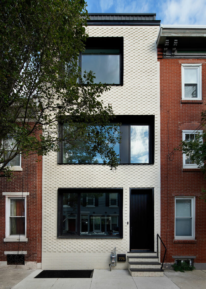 A modern, three-story townhouse with a white brick facade and large rectangular windows, nestled between two traditional red brick buildings. A tree partially obscures the left side of the house.