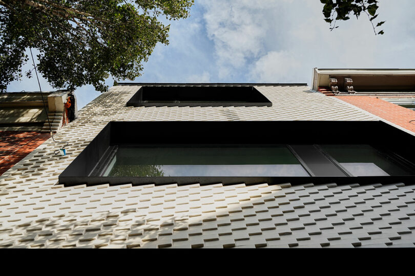 A low-angle view of a modern building facade adorned with interlocking white tiles and large black-framed windows, with trees and sky visible above.