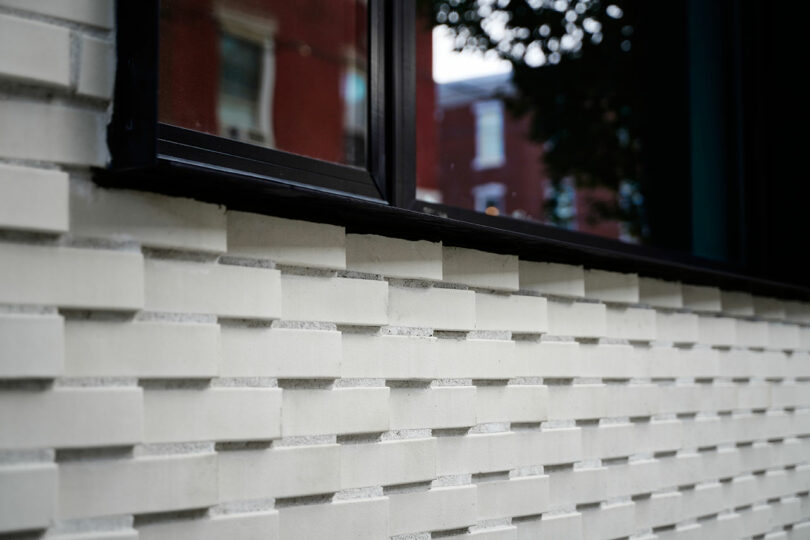 Close-up of a modern brick wall with a textured pattern and a window above it, reflecting surrounding buildings.