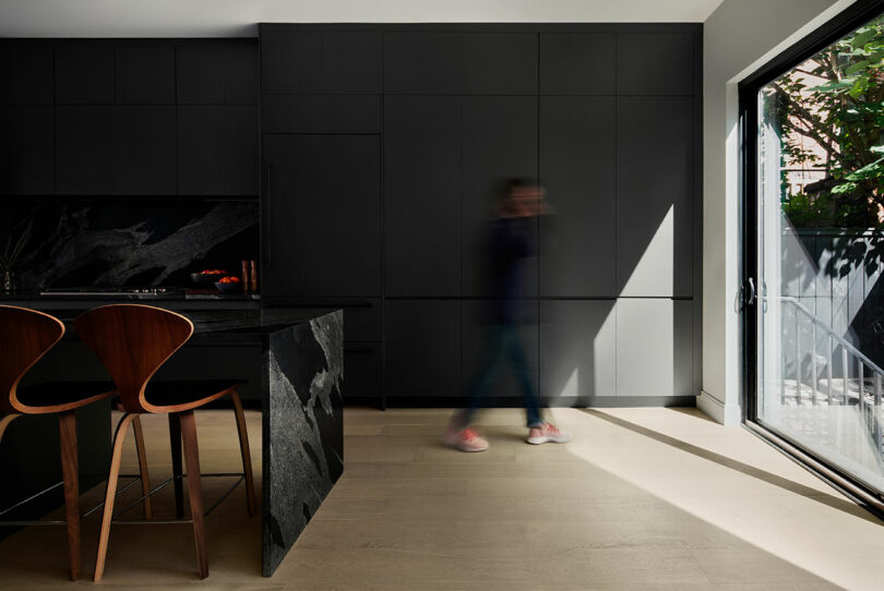 A person blurred while walking in a modern kitchen with black cabinets, wooden bar stools, and a large glass door letting in natural light.