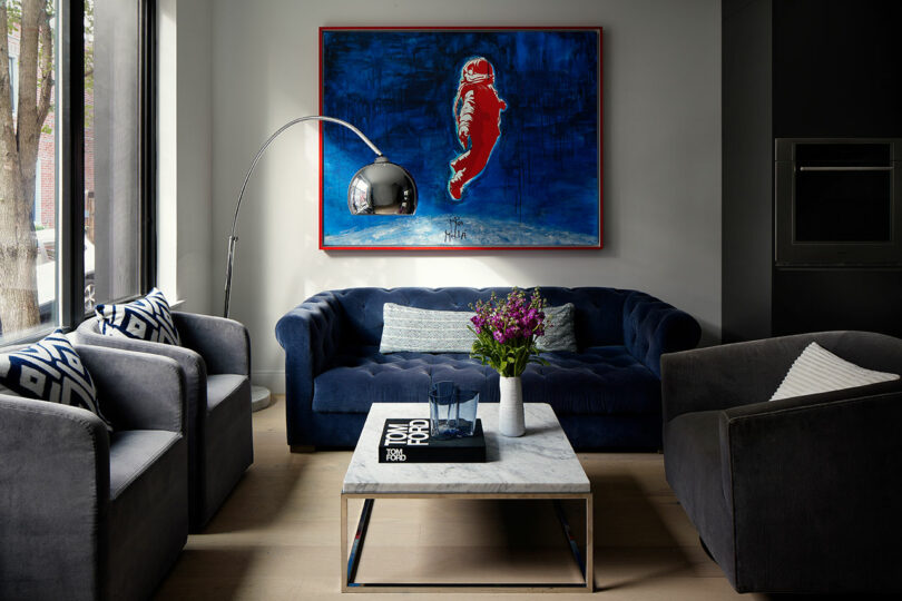 Modern living room with a dark blue sofa, two grey armchairs, a marble coffee table, and a large painting of a figure on the wall. A floor lamp arcs over the sofa, and a vase of flowers decorates the table.