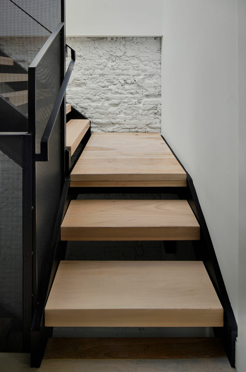 A modern staircase with light wooden steps, black metal frame, and handrail is positioned next to a white brick wall.